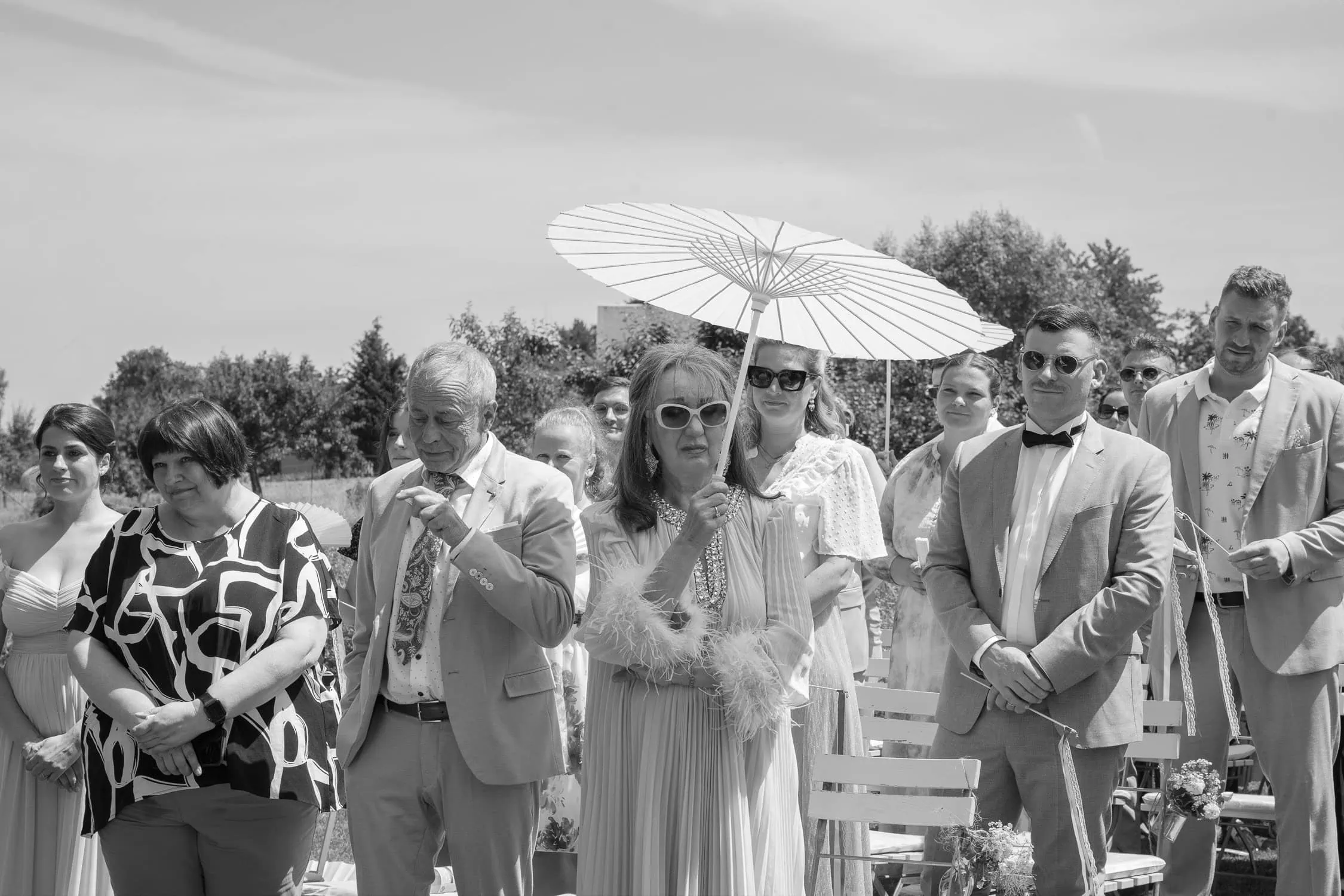 Gäste bei der freien Trauung in Schwarz-Weiß, eine Frau trägt eine Sonnenbrille unter einem Schirm - natürliche Hochzeitsfotografie